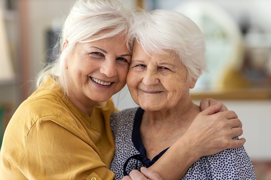 A smiling older woman hugs her caregiver in the front yard. 