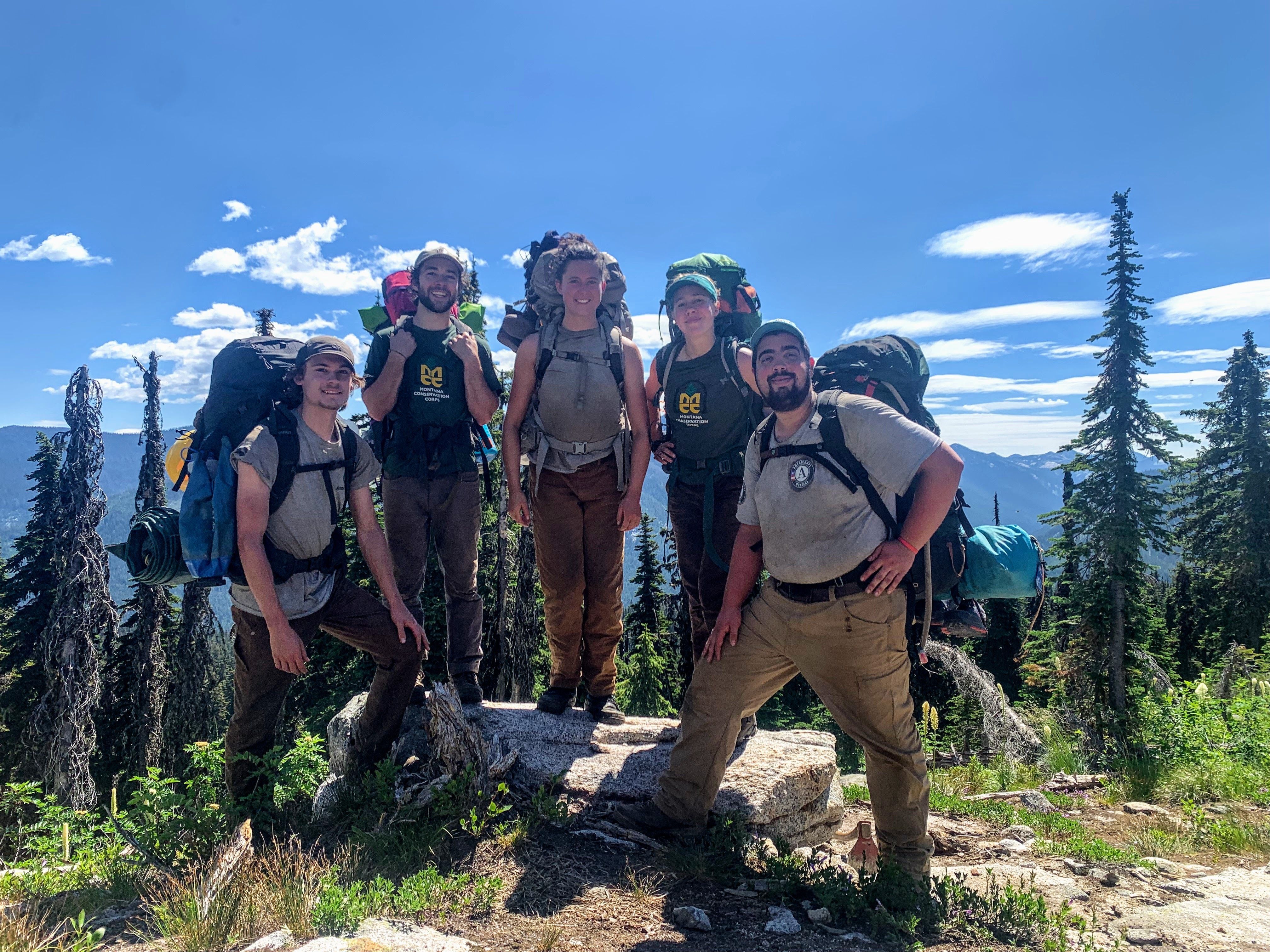 A smiling crew poses standing around and on a rock. They are all wearing backpacks.