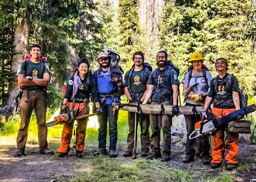 [Image Description: Seven MCC Members on a Saw crew, all standing with their covered chainsaws and chaps in a deep forest.]