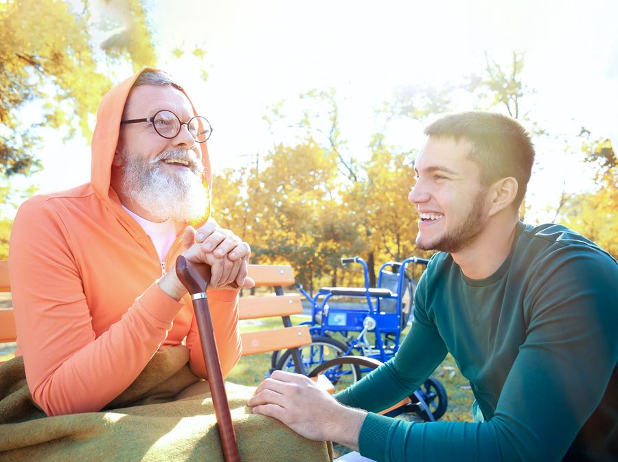 Smiling older man with cane is sitting on a park bench in front of his wheelchair. A young man crouches in front of him and is also smiling.