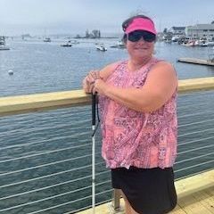 A smiling woman wears a hat and sunglasses. She is standing with her white cane. Water is behind her.