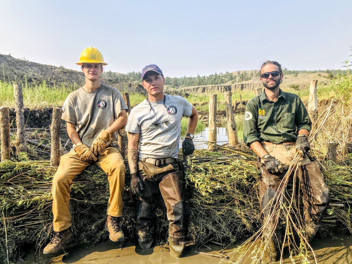 [Image Description: Three MCC members sit on the beaver dam analog they created, wearing their MCC uniforms and smiling.]