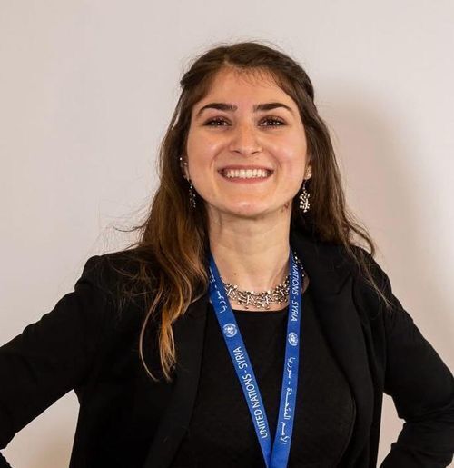 A woman with dark hair smiles broadly at the camera. She is dressed in black with a blue name tag around her neck. The name tag says United Nations, Syria