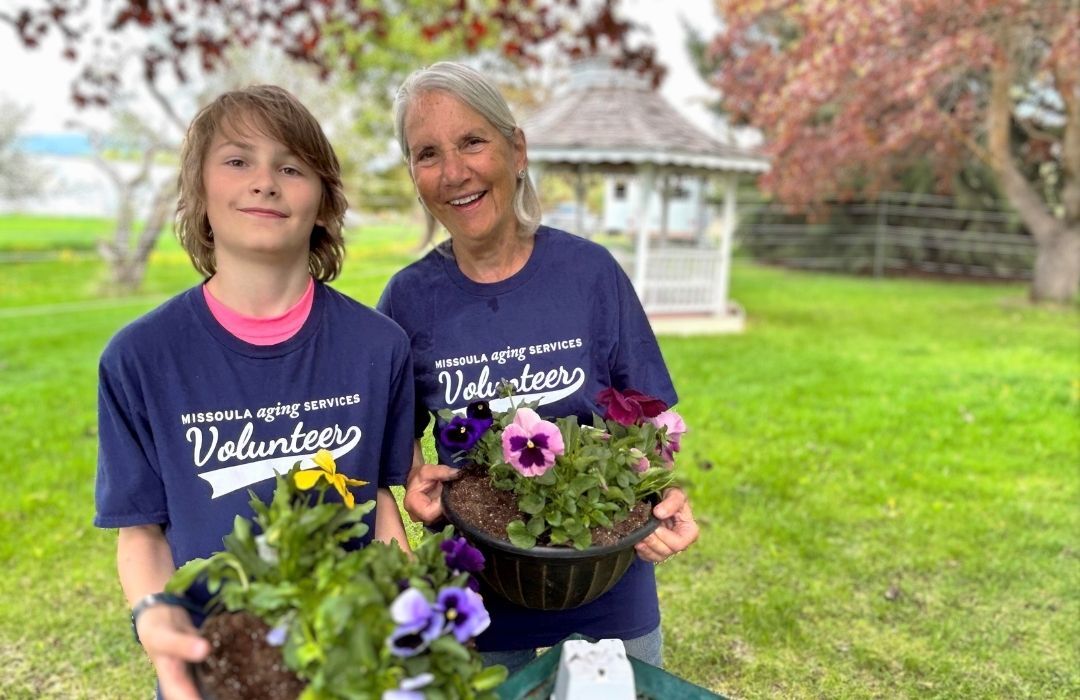 Smiling male and female volunteers wearing their Senior Corps t-shirts at a playground. They're holding shovels and standing in front of a wheelbarrow.