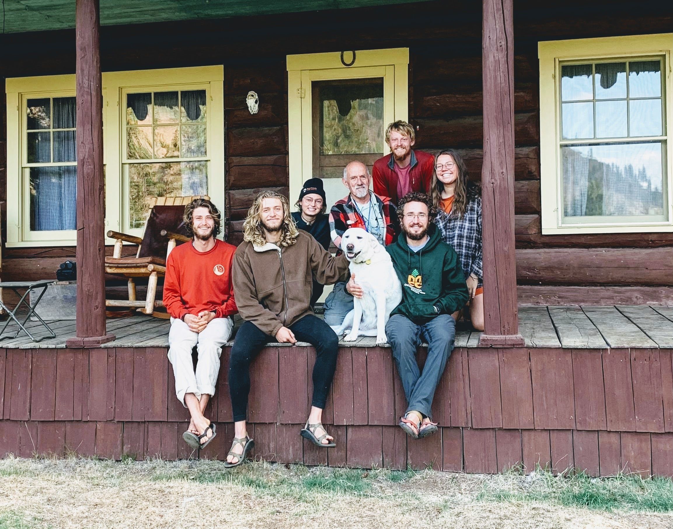 A group sits on a porch of a cabin, smiling. There is a yellow lab.