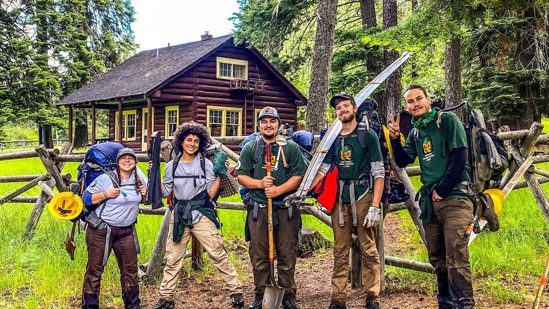 [Image Description: Five MCC members stand together, in front of a rustic cabin. One of the members has a crosscut saw resting on their shoulder. They all have their loaded backpacks on, and look ready to hit the trail!]
