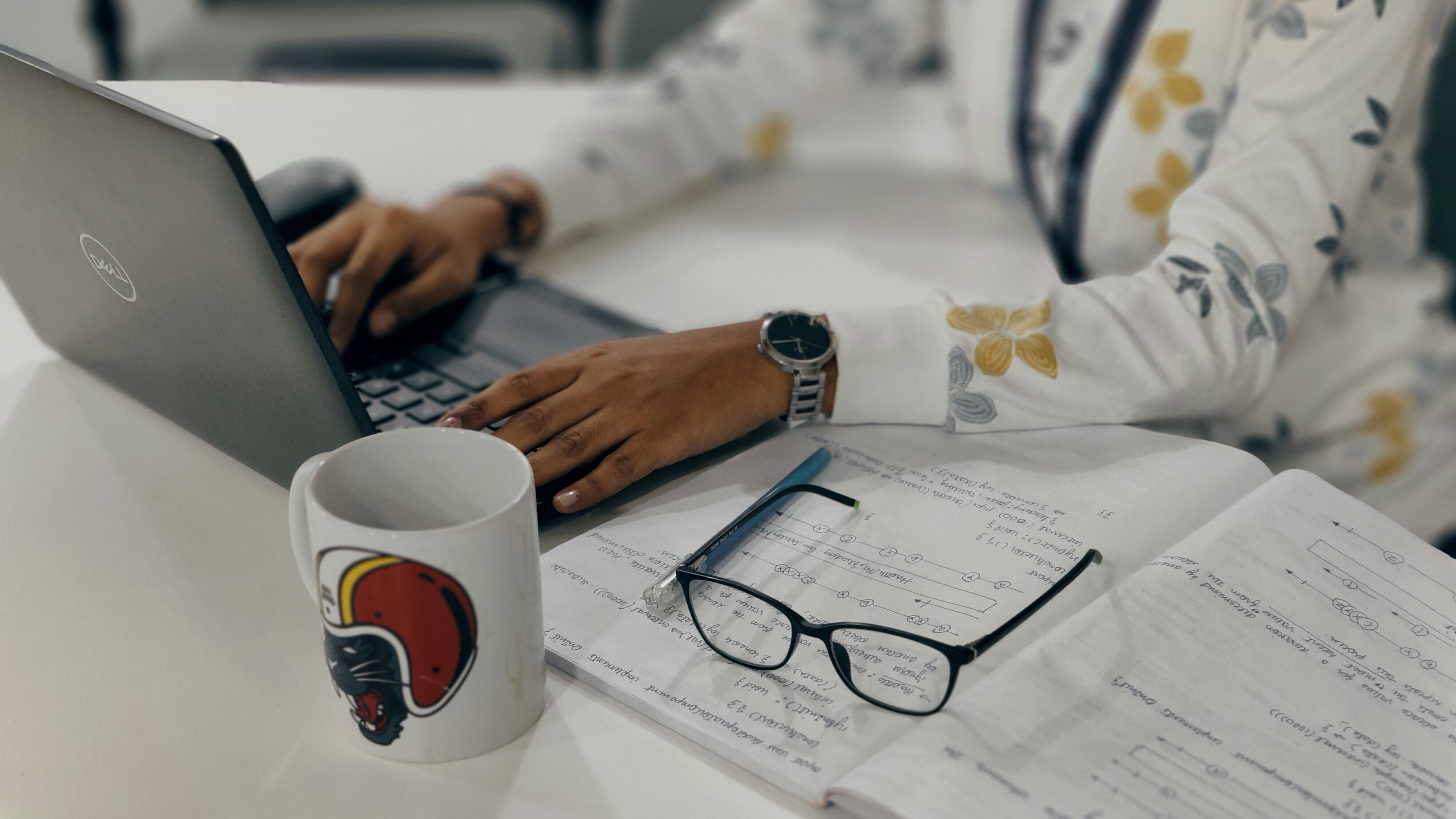 Woman using laptop with notebook, glasses, and mug on desk