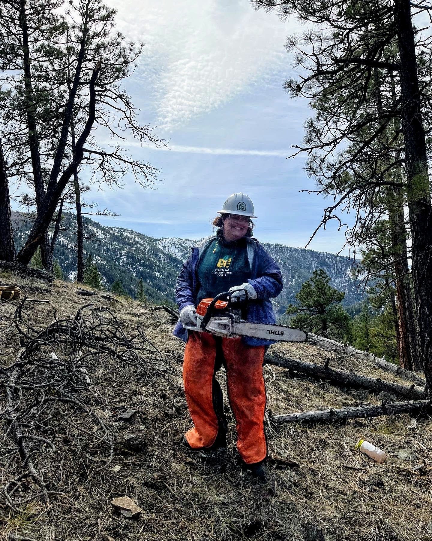 A youth leader strands in a clearing surrounded by trees, with snow capped mountains in the background. They are holding a chainsaw while wearing chaps and a helmet.