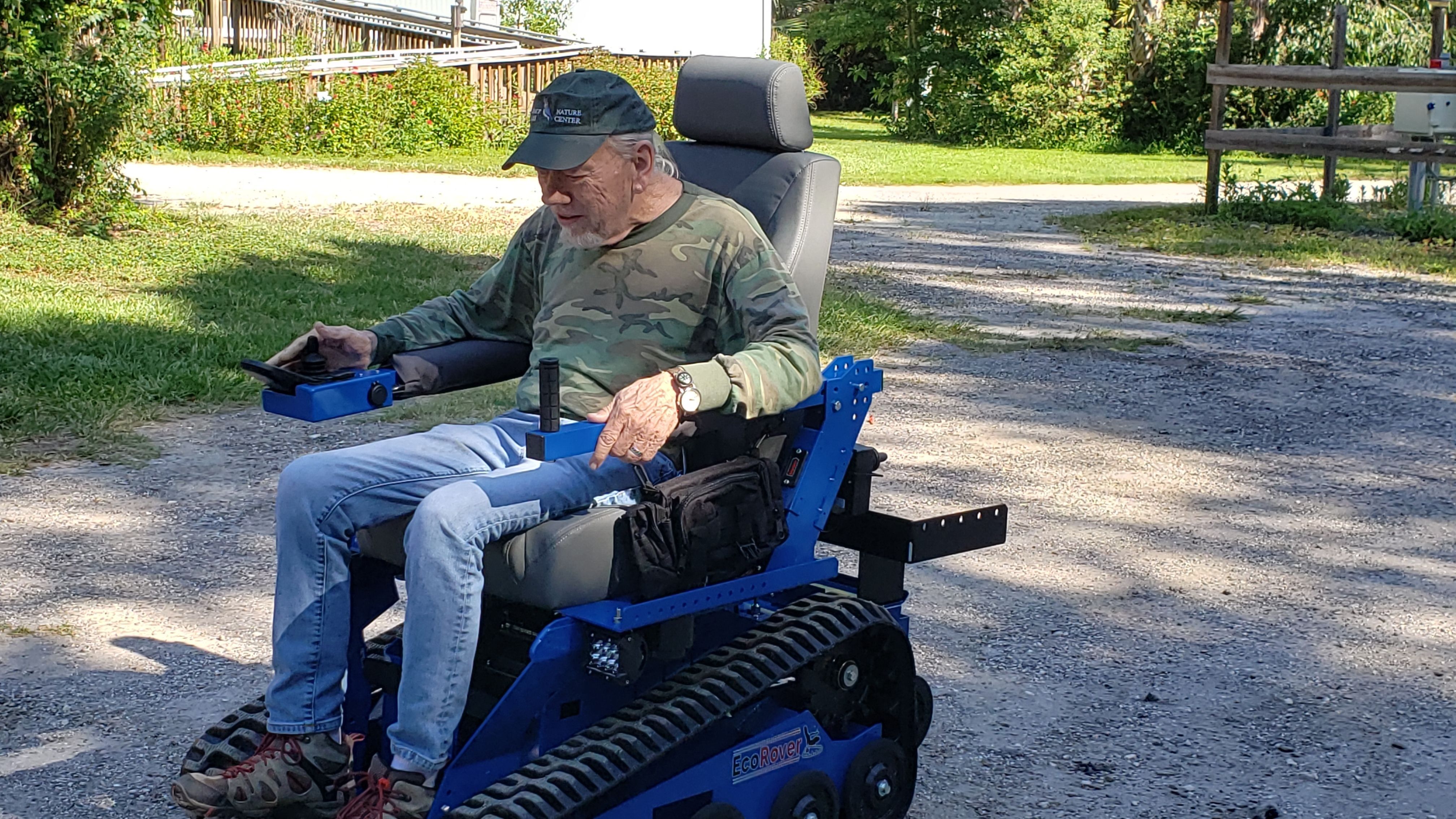 An older gentleman dressed in blue jean, camo shirt and ball cap is sitting in the blue EcoRover chair with joy stick in hand and tracks for wheels. 
