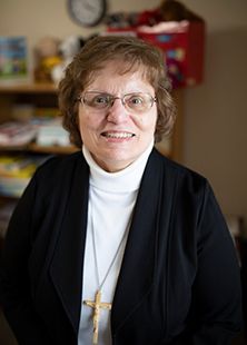 Sister Fredrica wears a white turtleneck and black sweater. She is smiling at the camera. 