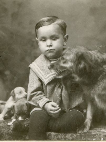 A portrait style black & white photograph of a young boy sitting with a golden retriever dog and two young puppies.  The boy is wearing his Sunday best clothes.