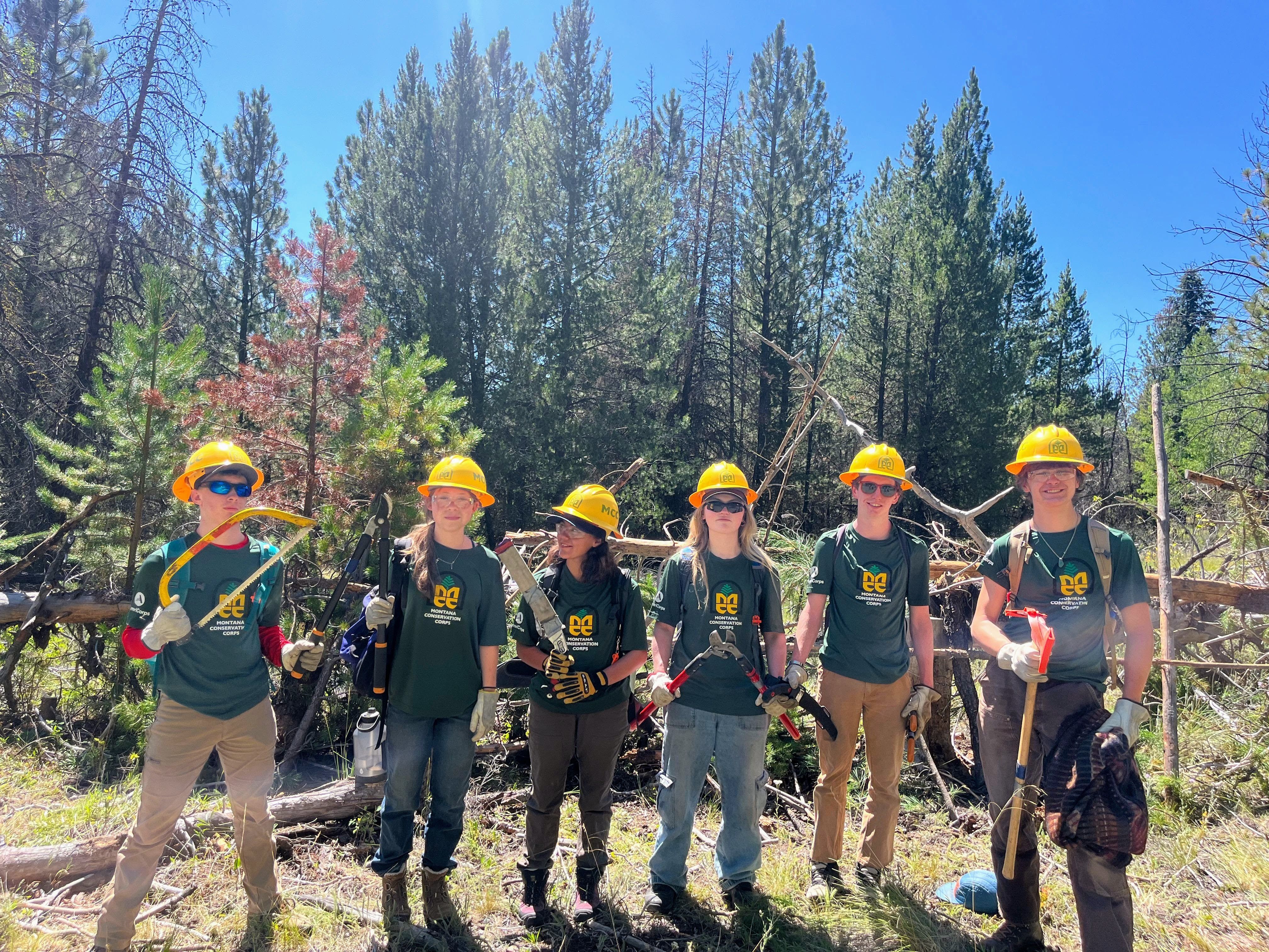 [Image Description: Six MCC members standing together on a trail, overlooking a mountainous landscape, surrounded by green shrubs and pine trees.]