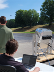 Two men, a laptop, and a large metallic device outdoors with a hovering drone in the background.