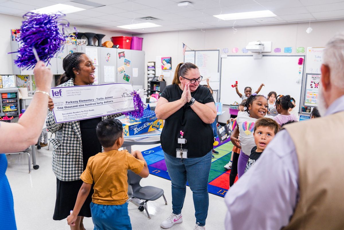 lef volunteer surprising a teacher with a big presentation check while children scream in excitement