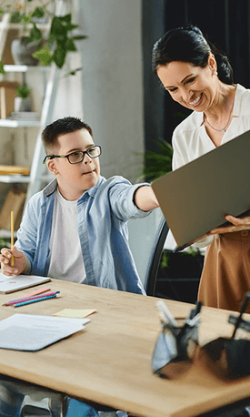 A young boy with Down syndrome seated at a desk reaches toward a smiling woman holding a laptop in a bright room with pencils and papers on the desk