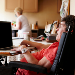 Woman in a wheelcair working on her laptop while her personal care assistant works in the kitchen.