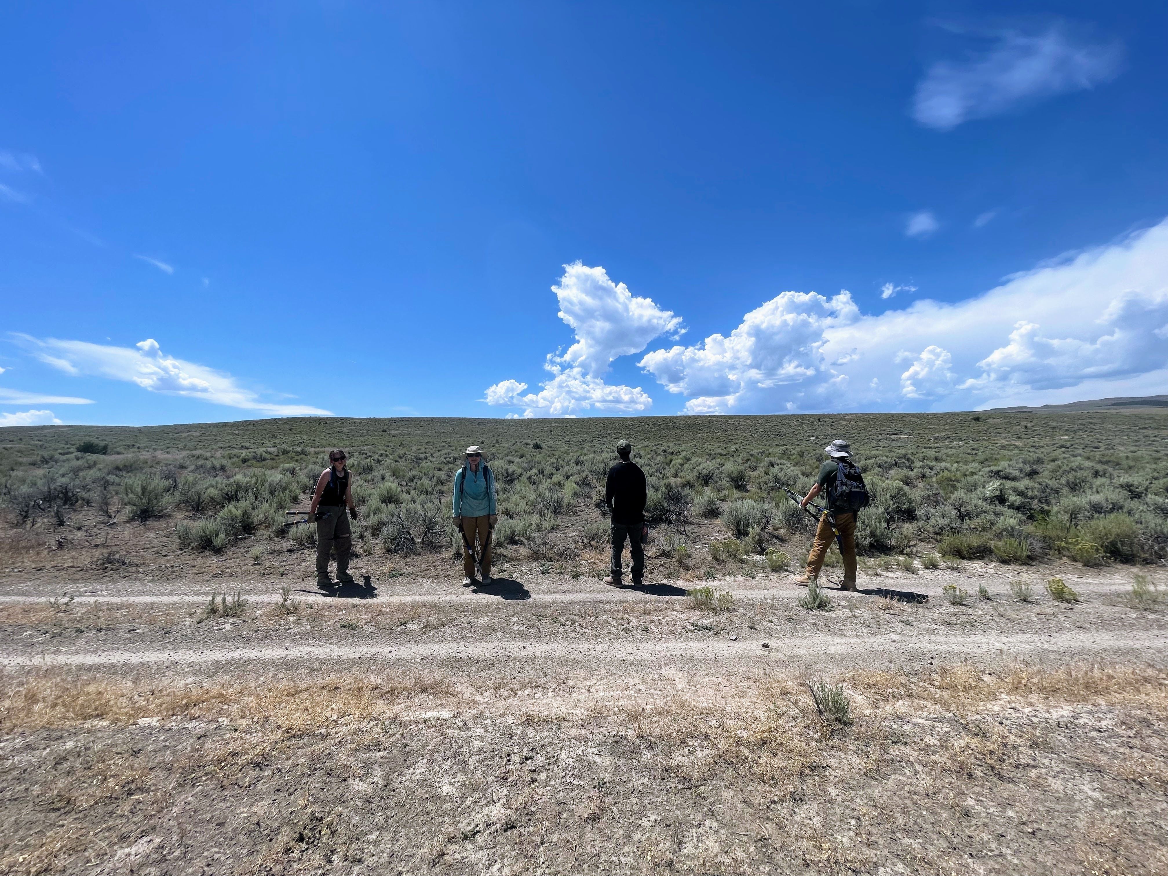 A crew stands on a dirt road in the desert with a bright blue sky and sagebrush behind him.