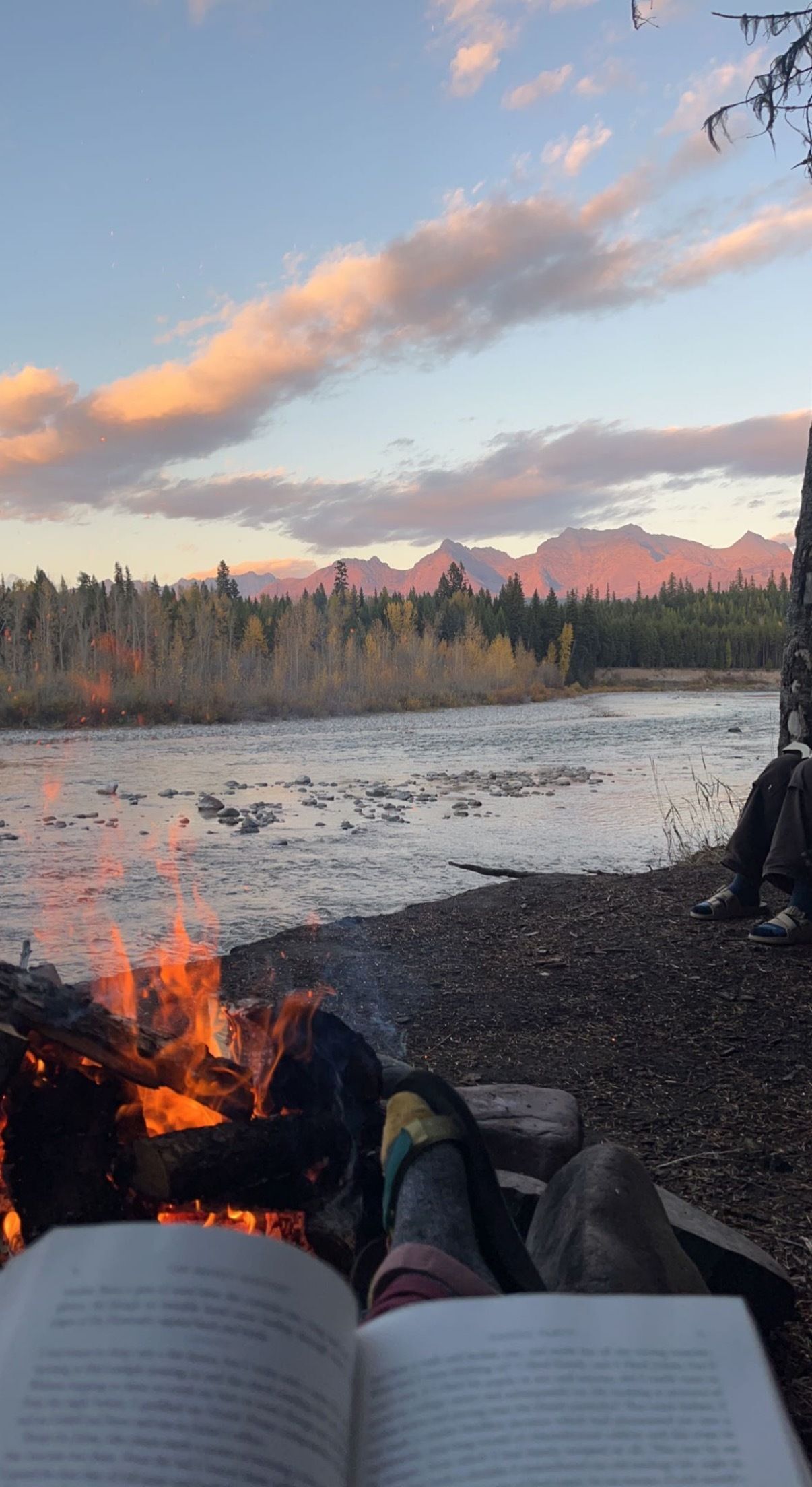 A person sits on a stream bank, with a burning campfire in front of them and mountains in the distance.