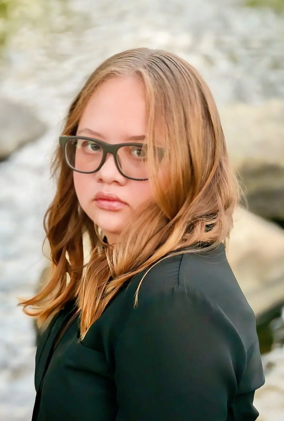 Zeda is a white woman with Down syndrome and dark blonde hair. She wears dark clothes and dark-rimmed glasses. Her body faces to the left, with her head turned forward. Her face smolders.