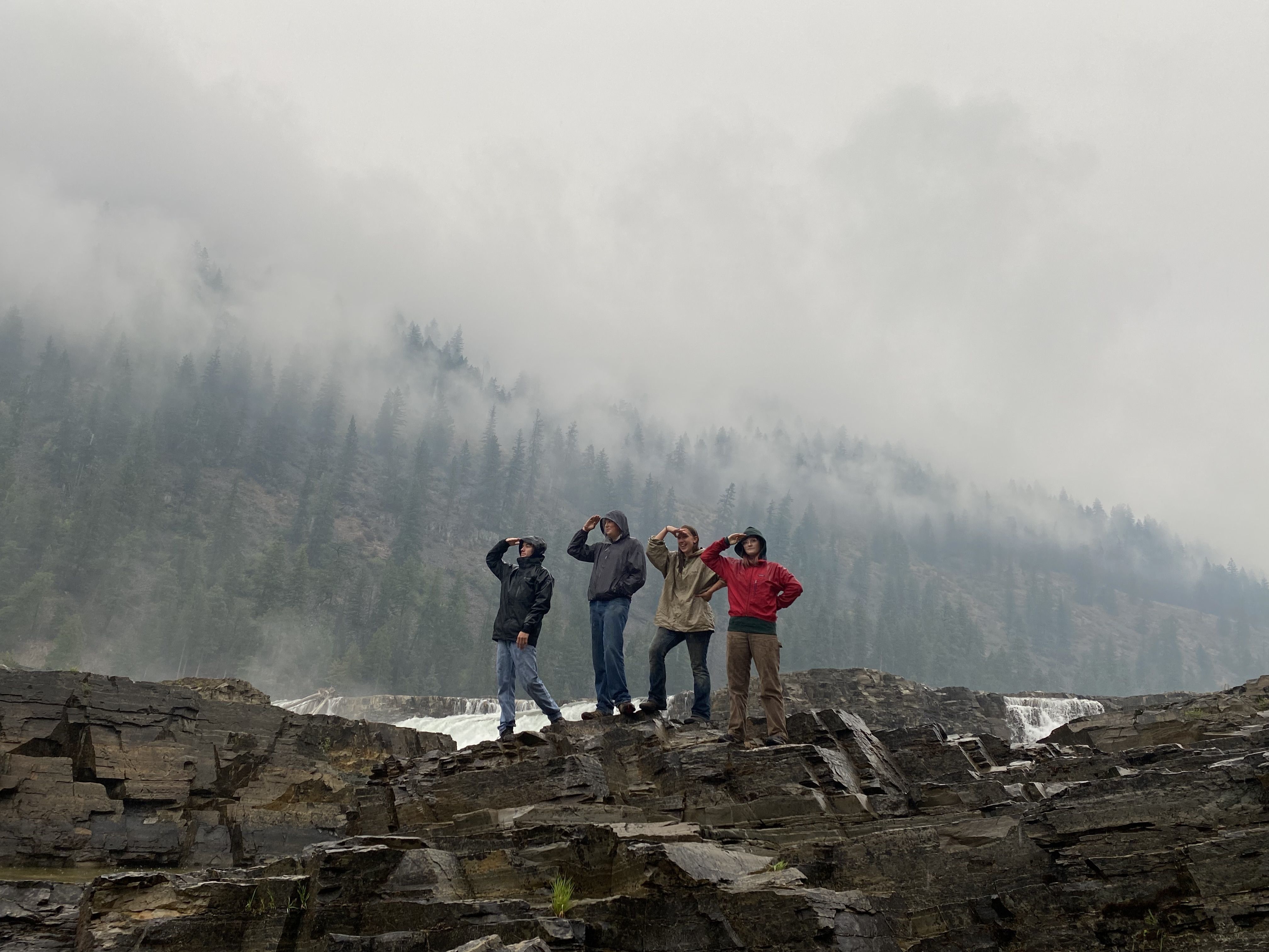 A crew stands on some rocks looking to the left, in front of a misty, forested hillside