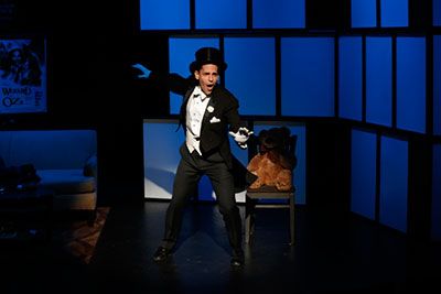 ACCORDING TO GOLDMAN - 2013. Stephen Drabicki is wearing a tuxedo with white gloves and a black top hat. He is a magician and he is performing a magic trick in a dim lighting. 