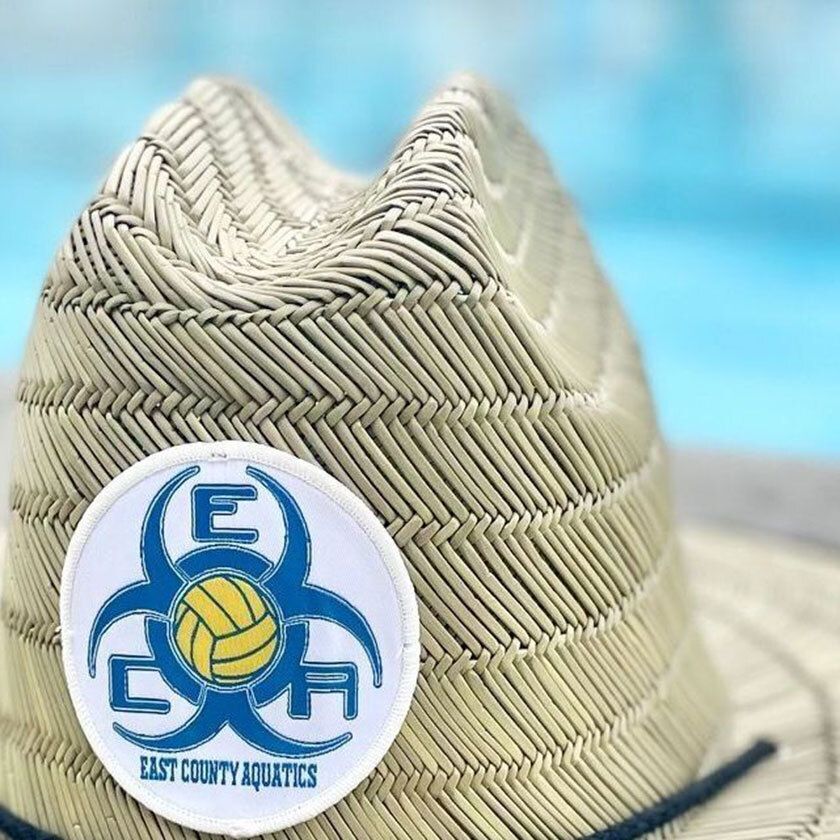 Close-up of a straw hat with a circular patch displaying a biohazard symbol and text "EAST COUNTY AQUATICS."