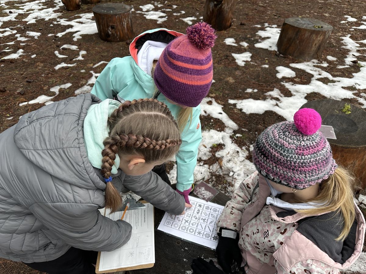 Three young girls dressed in winter gear studying snow crystals on a page and drawing them on their clipboard.
