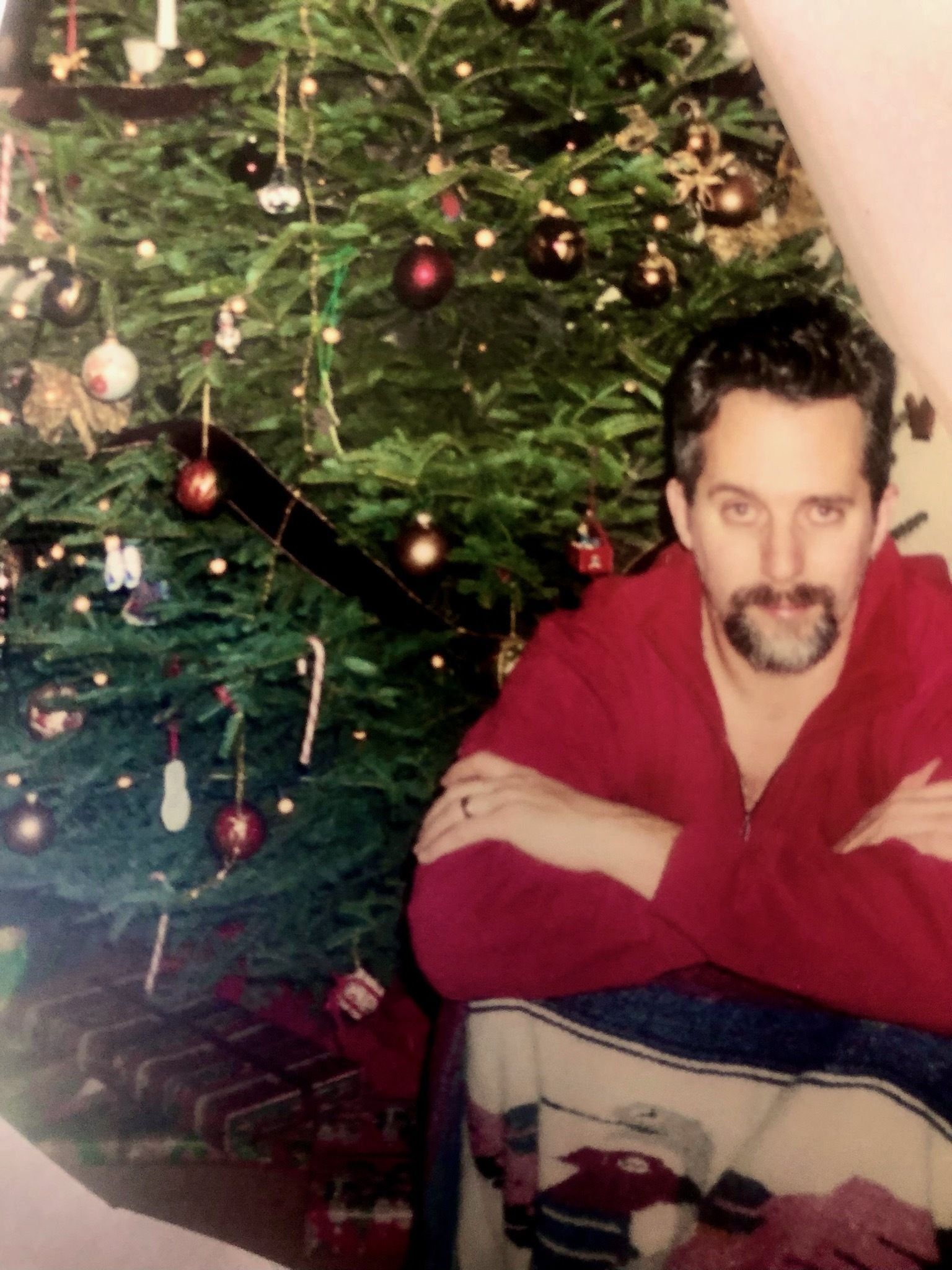 White man poses next to a Christmas tree in a red shirt.
