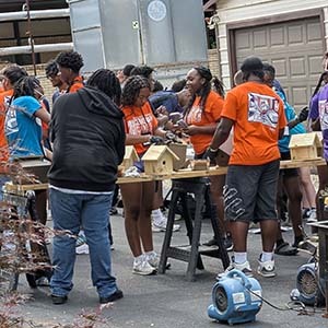 Group of volunteers in orange and purple shirts building birdhouses at tables in a driveway
