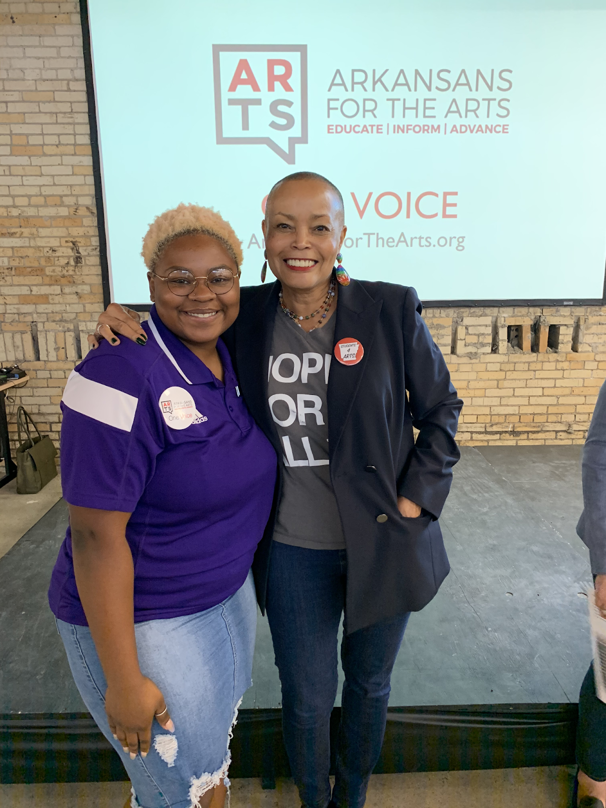 A college students in purple UCA shirt poses with state senator Joyce Elliot in front of a projector screen with the Arkansans for the Arts logo displayed prominently.