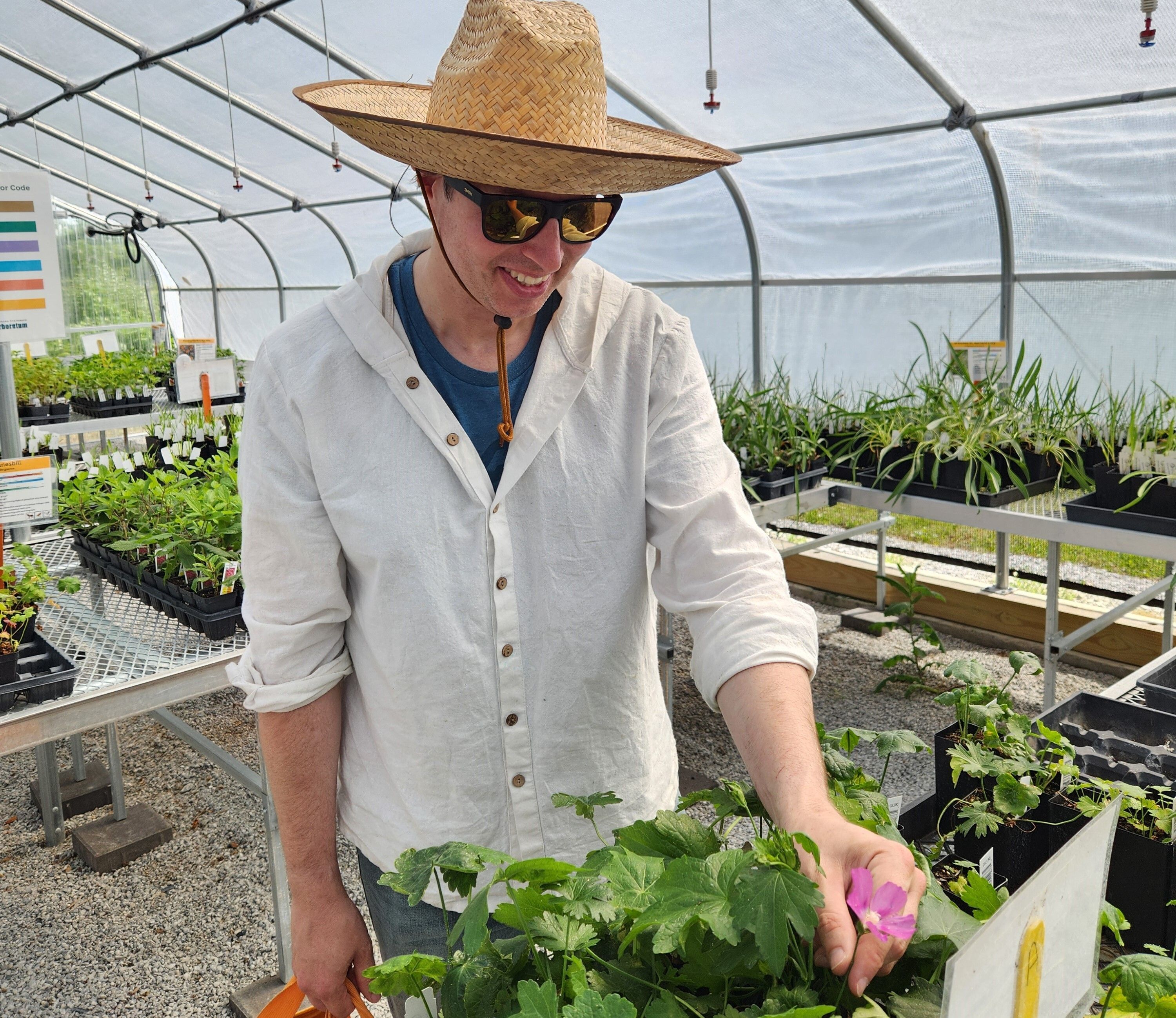 Man wearing a white shirt and a straw hat shops for plants inside a greenhouse. 