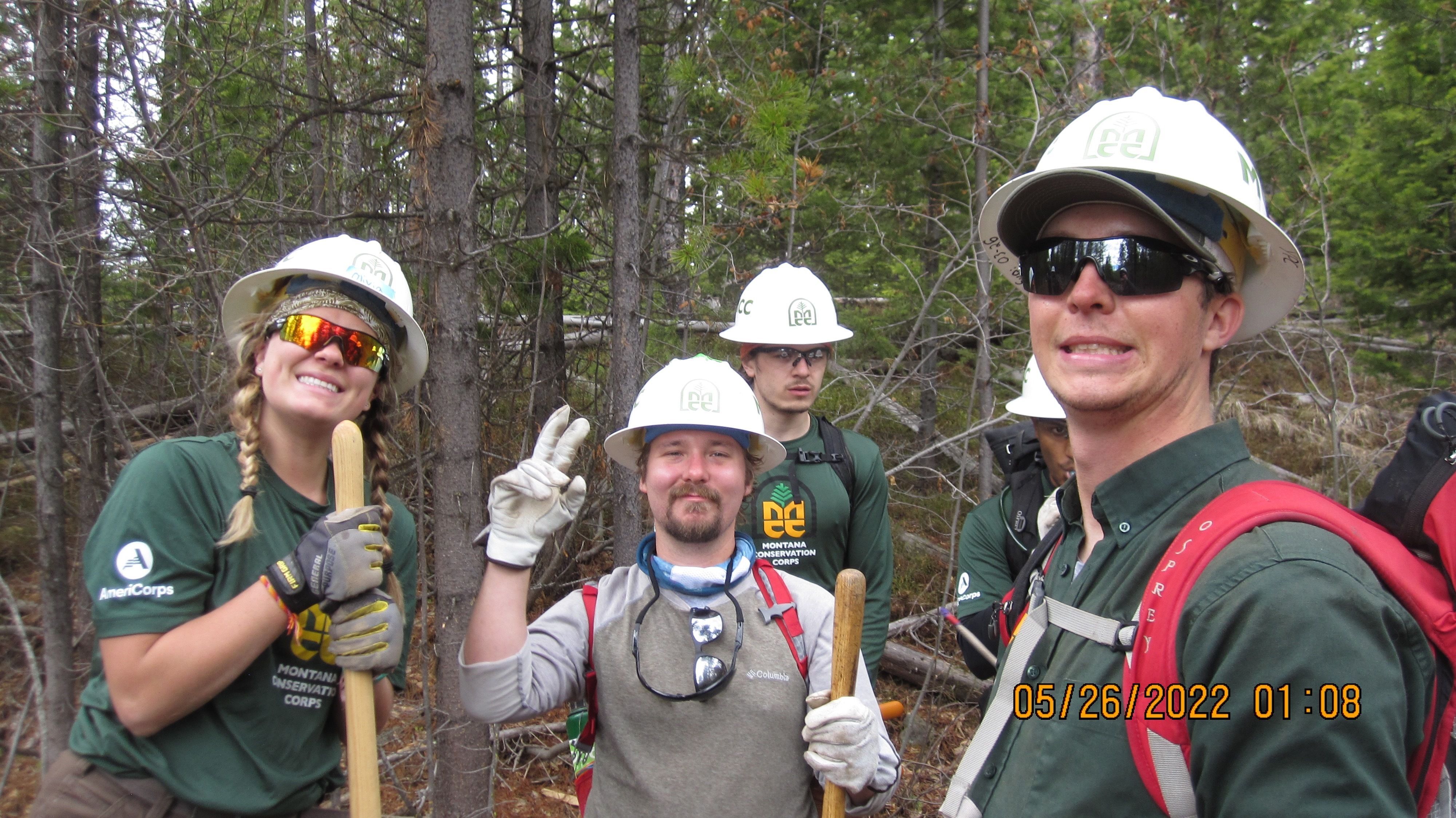 A group of people wearing white helmets, youth leaders, make silly faces while posing for the camera. There are trees in the background.