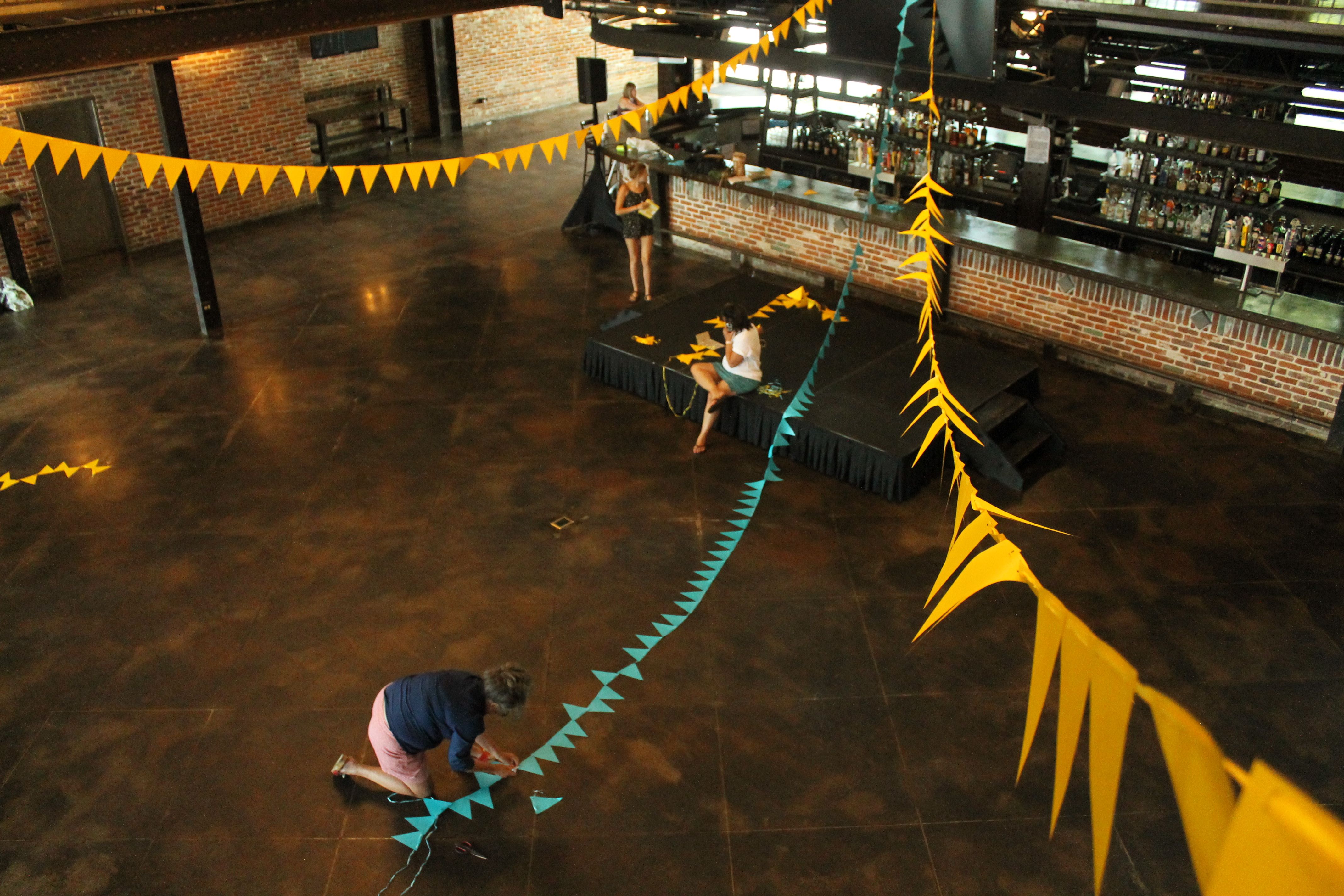 Woman kneels on ground; she is putting together yellow and turquoise flags, some of which hang from the ceiling above her