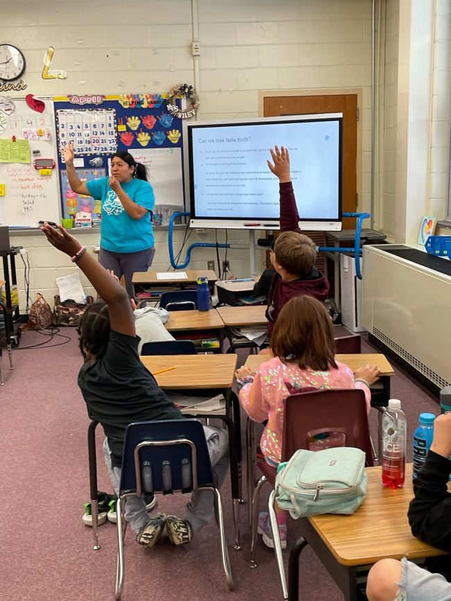 We are facing the backs of third graders seated at their desks in the foreground in the Morrisville public schools. The students are stretching up to raise their hands to answer nutrition questions posed by Odalis Macario in the back of the picture facing