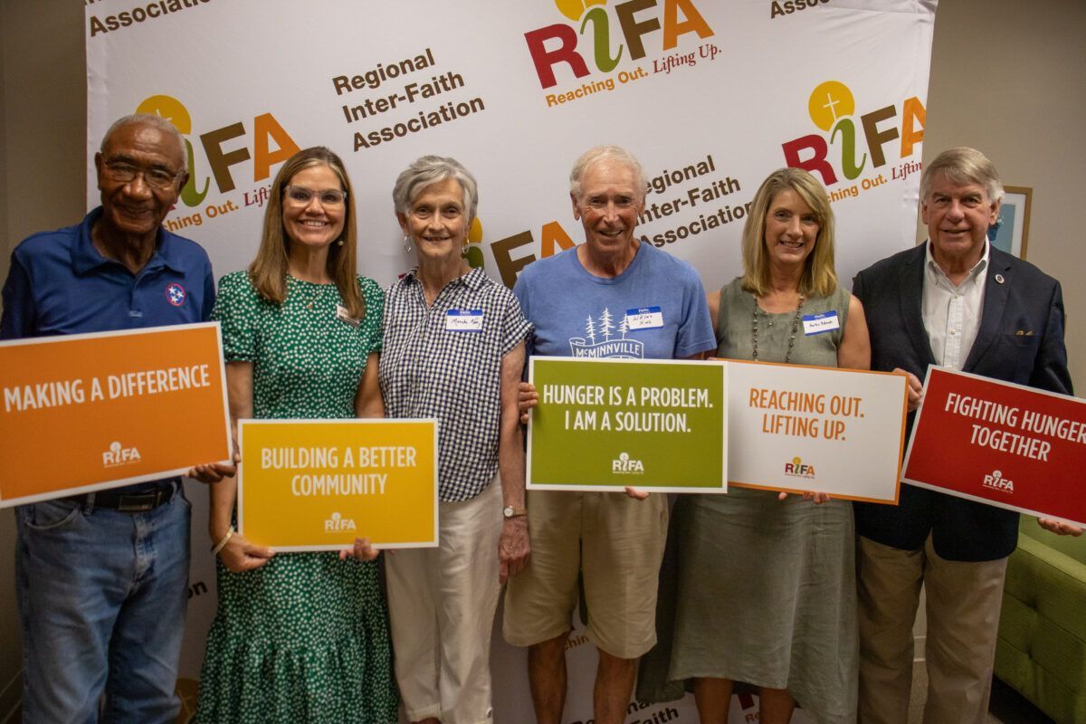 Six people holding signs with messages about community and hunger relief in front of a Regional Inter-Faith Association backdrop.