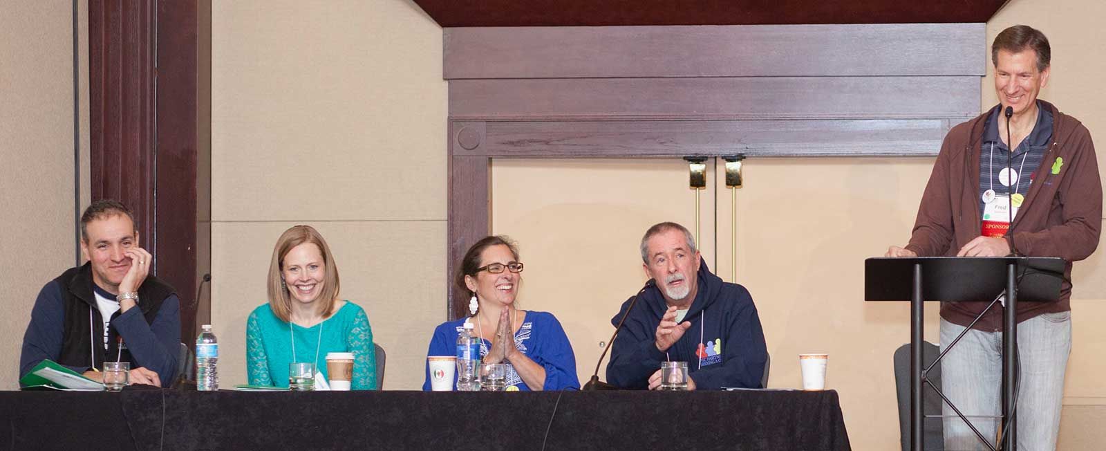 A man stands smiling behind a podium. Beside him is a long table with a panel of four seated people, two women in the middle and a man on either end.