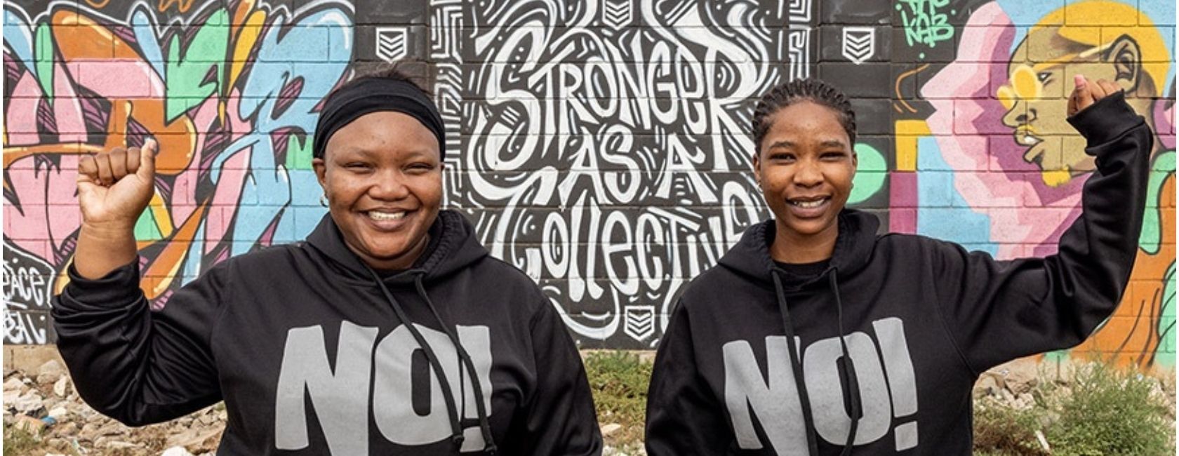 Two women with sweatshirts saying "No!" standing together, smiling, and raising their outside arms 