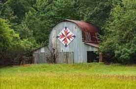 A weathered barn stands in the middle of a field of green grass. Hung prominently above the doors is a brightly colored quilt block of epic proportions.