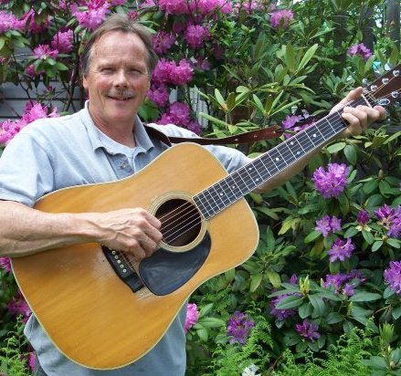 Man posing with a guitar in front of pink flowers