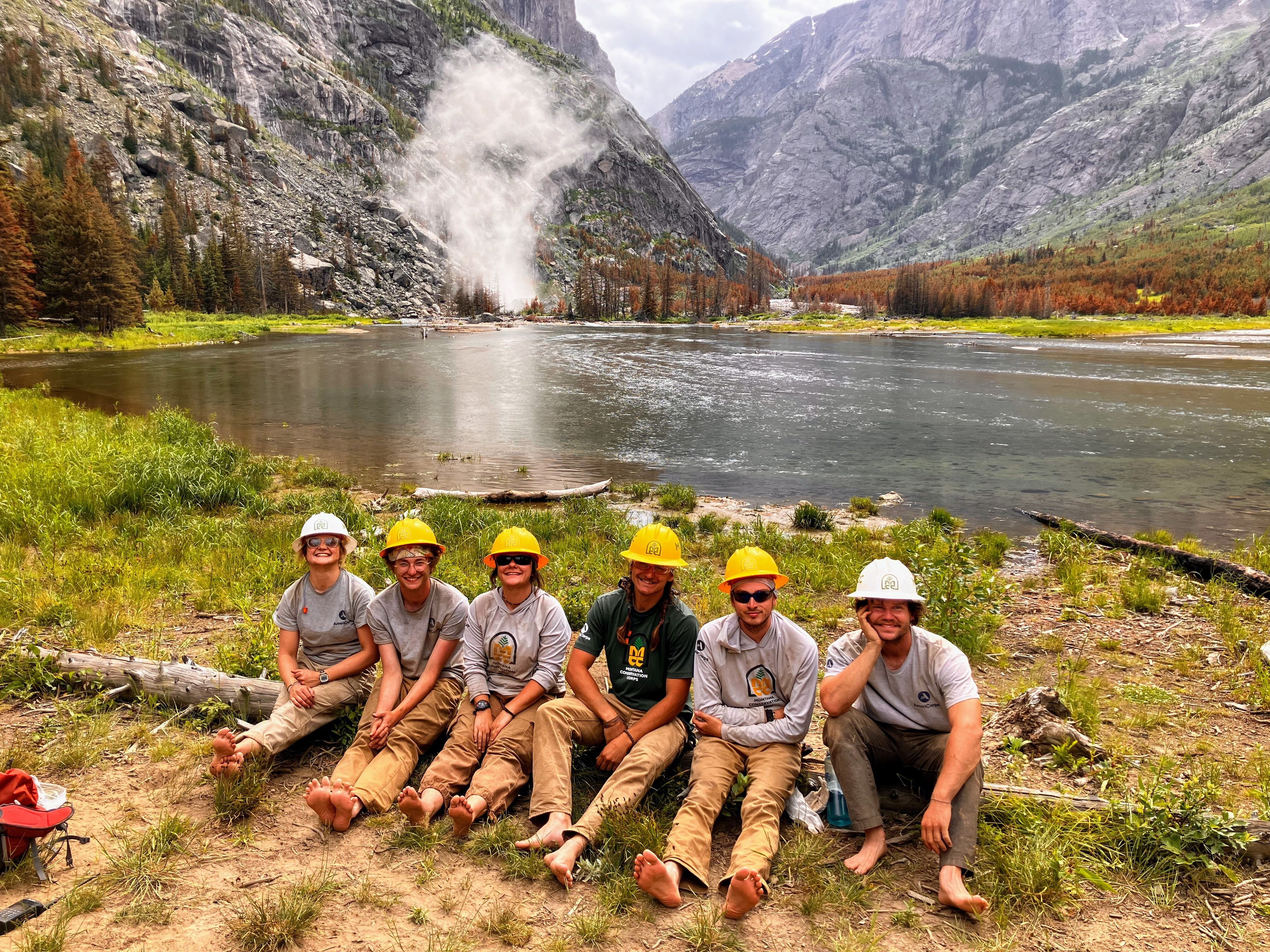 A crew sits on a log on the shore of a lake, with a cloud of dust in the background from an explosion.