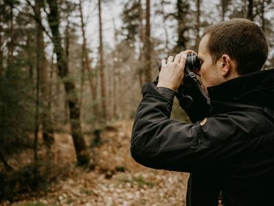 A person stands in the woods in the woods wearing a black jacket and looking through binoculars.