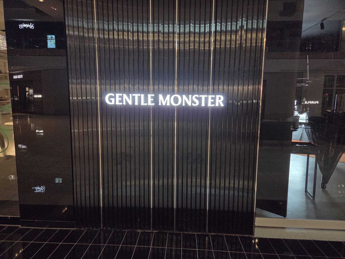 Illuminated “Gentle Monster” sign made from precision-cut acrylic and backlit with LED lighting, mounted on a vertical ribbed black wall inside a modern retail space.