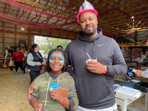 A girl and her father stand inside a large barn eating frozen refreshments. The girl is in bright blue face paint.