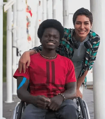 The image shows two smiling individuals outdoors near a row of flagpoles. One person is sitting in a wheelchair wearing a red shirt and a beaded necklace. The other person stands beside them with a hand on their shoulder wearing a plaid shirt