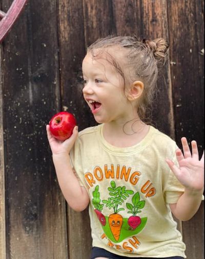 Young girl stands with her hands held up to her shoulders with an spple in her right hand.