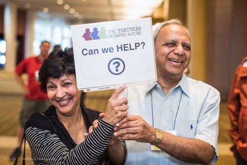 A smiling couple holding a sign that says Can We Help. Behind them are other conference goers.