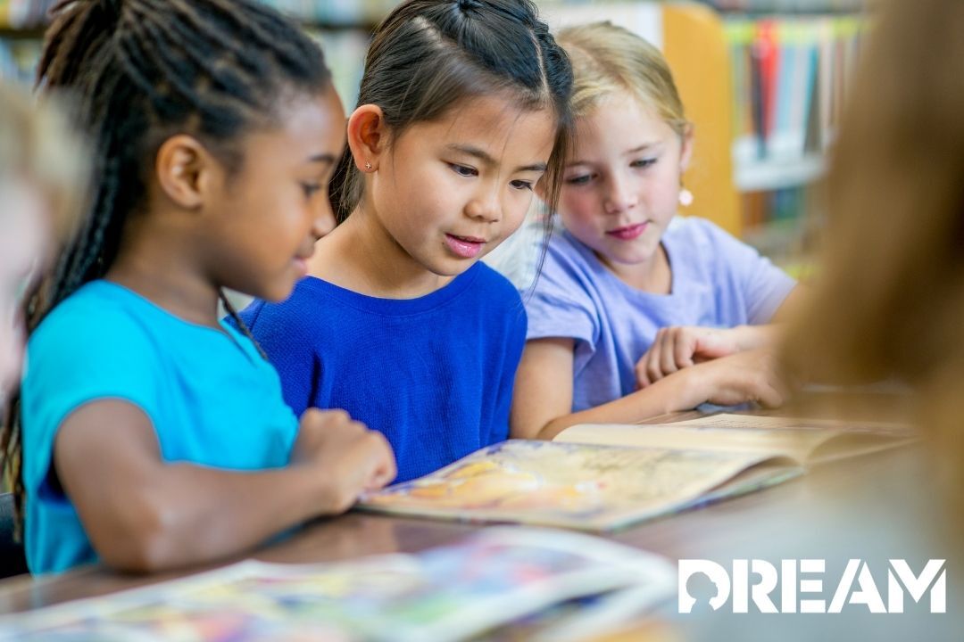 Three young kids are sitting at a table reading the same book and smiling