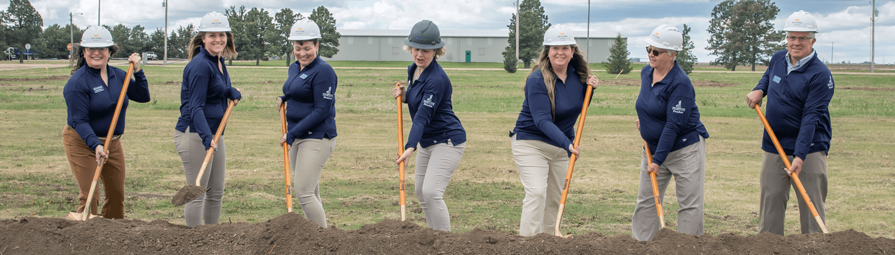 Seven people holding shoves, wearing construction hard hat, all wearing navy blue long sleeve tops with tan pants.