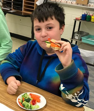 A student in a blue fleece sweatshirt with a hood looks up at the camera in the Morrisville, PA, school district enjoys a healthy sandwich made from humous and fresh organic vegetables, courtesy of the Snipes Farm & Education Center.