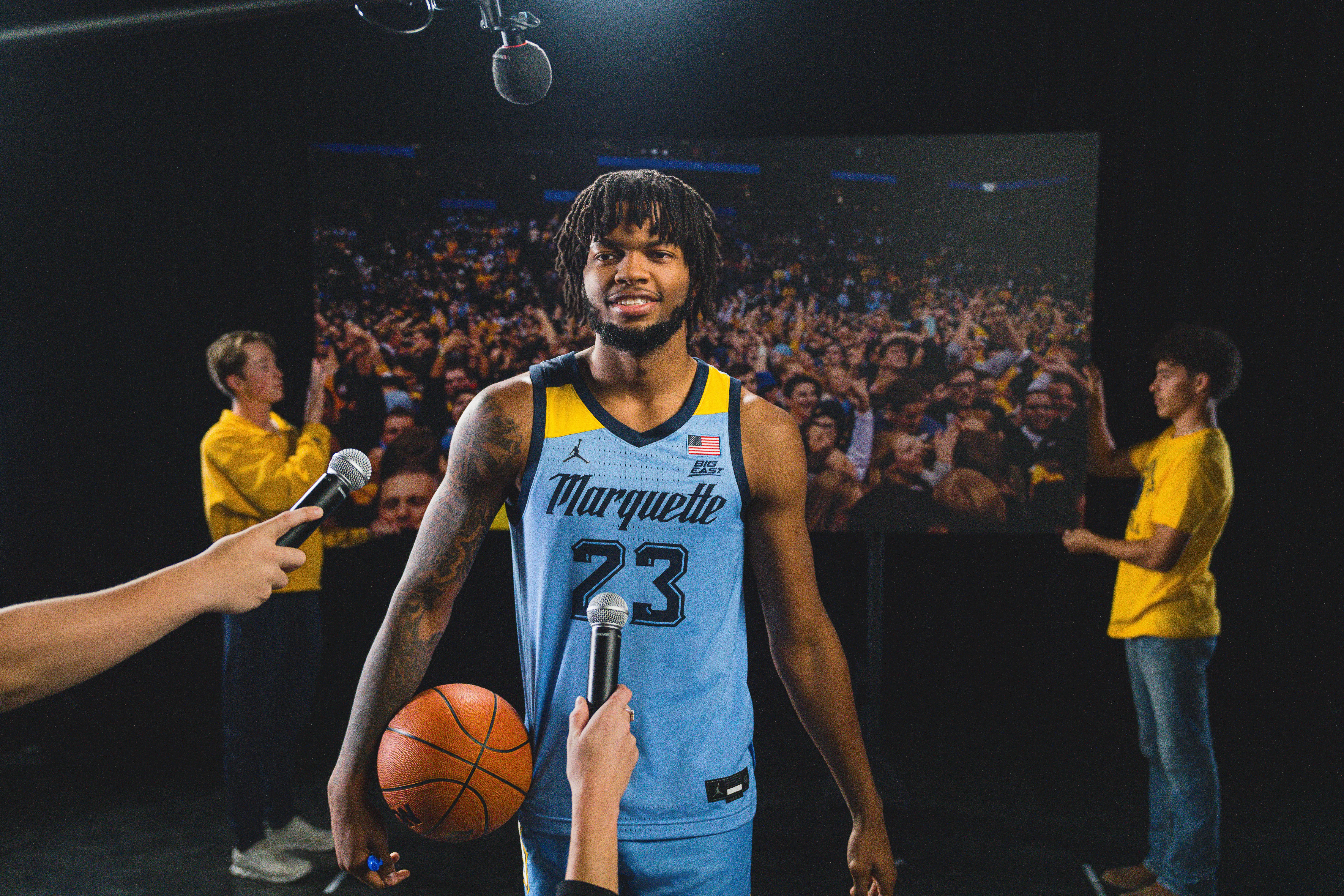 MU Basketball Player David Joplin is standing in front of a banner of fans. He is holding a basketball and has microphones in front of him. 